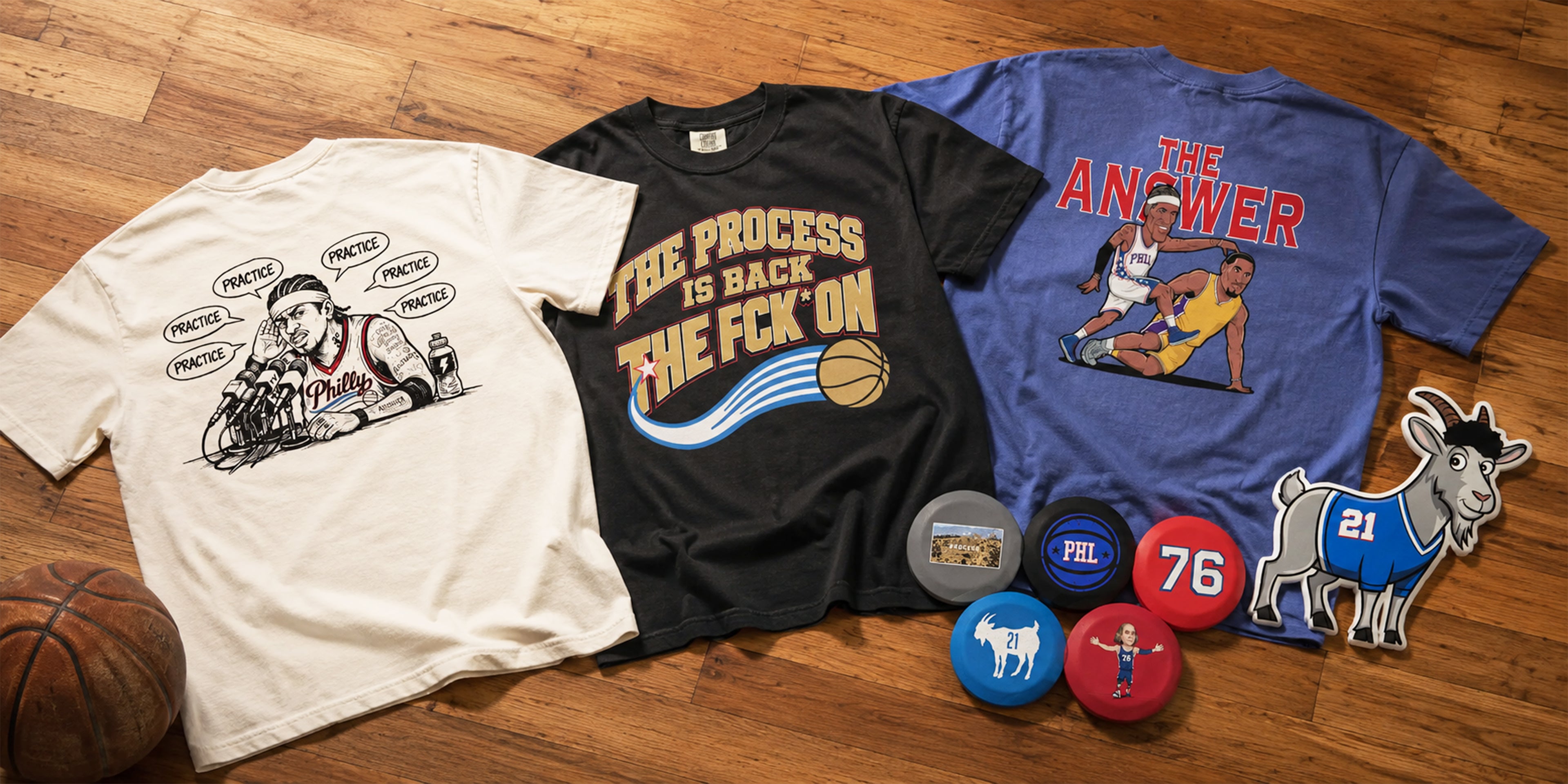 Three graphic T-shirts laid out on a wooden floor, along with several circular pins and a die-cut patch. The shirts feature basketball-themed graphics and text, including 'THE PROCESS IS BACK THE FCK ON' on a black shirt and 'THE ANSWER' on a blue shirt.